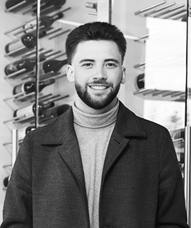 The finance manager of a luxury car dealership smiles at the camera. He is standing in front of a wine display.